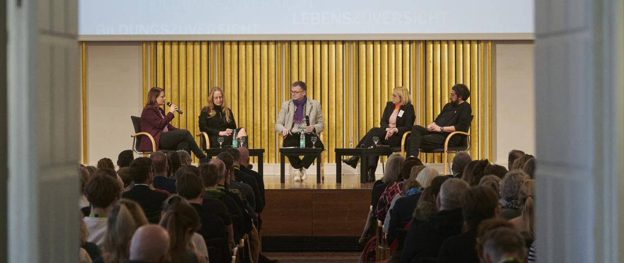 Fünf Personen sitzen auf einer Bühne in einem Saal vor Publikum.