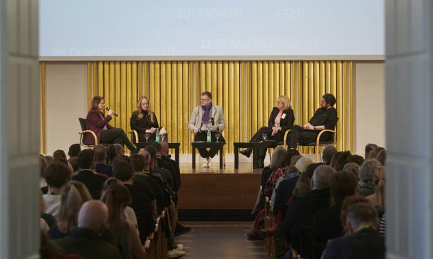 Fünf Personen sitzen auf einer Bühne in einem Saal vor Publikum.